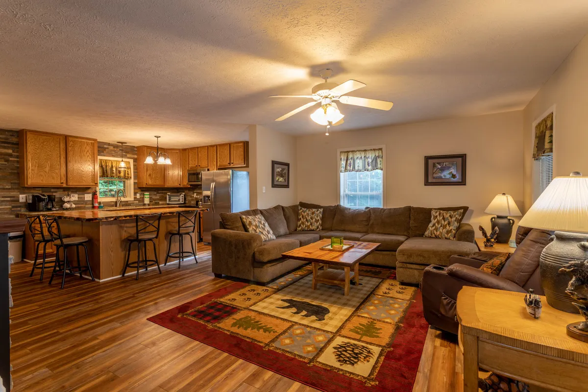 A wide view of the main living area, showcasing a spacious, inviting sectional sofa, a large wooden coffee table, a decorative area rug, and a glimpse of the kitchen island in the background. The space is bathed in natural light, creating a warm and communal atmosphere.