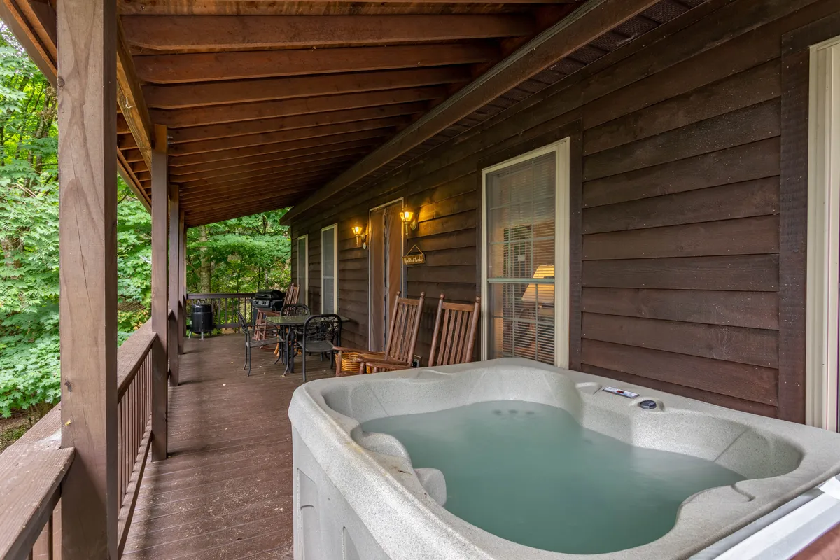 The image features a cozy wooden porch with a hot tub and several rocking chairs. The setting emphasizes a peaceful retreat, with the hot tub well-positioned for enjoying sunset or starlit views. The natural wood tones and soft lighting evoke relaxation and connection with the outdoors.