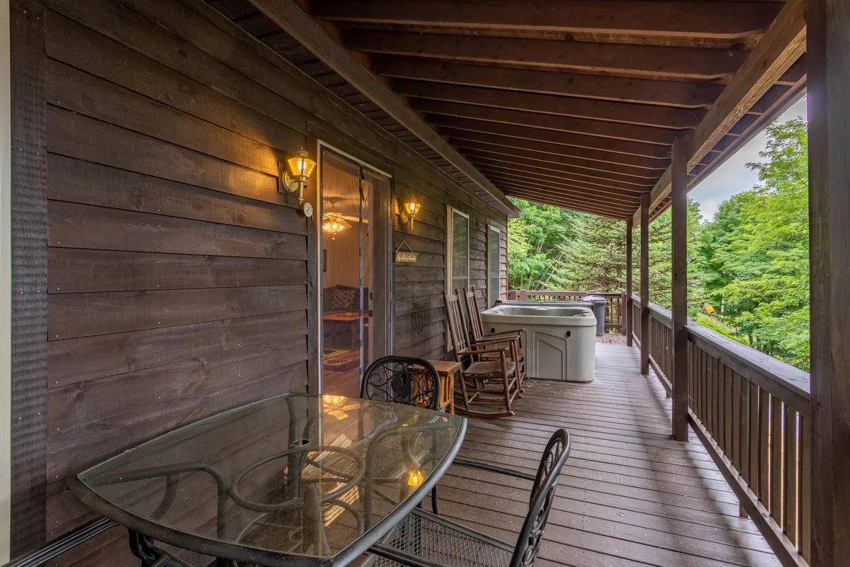 This exterior image showcases a private deck furnished with a glass table, inviting rocking chairs, a prominent hot tub, and a rustic wooden railing. Lush greenery surrounds the space, creating a tranquil atmosphere perfect for soaking in nature. The deck appears spacious and designed for comfort and relaxation.