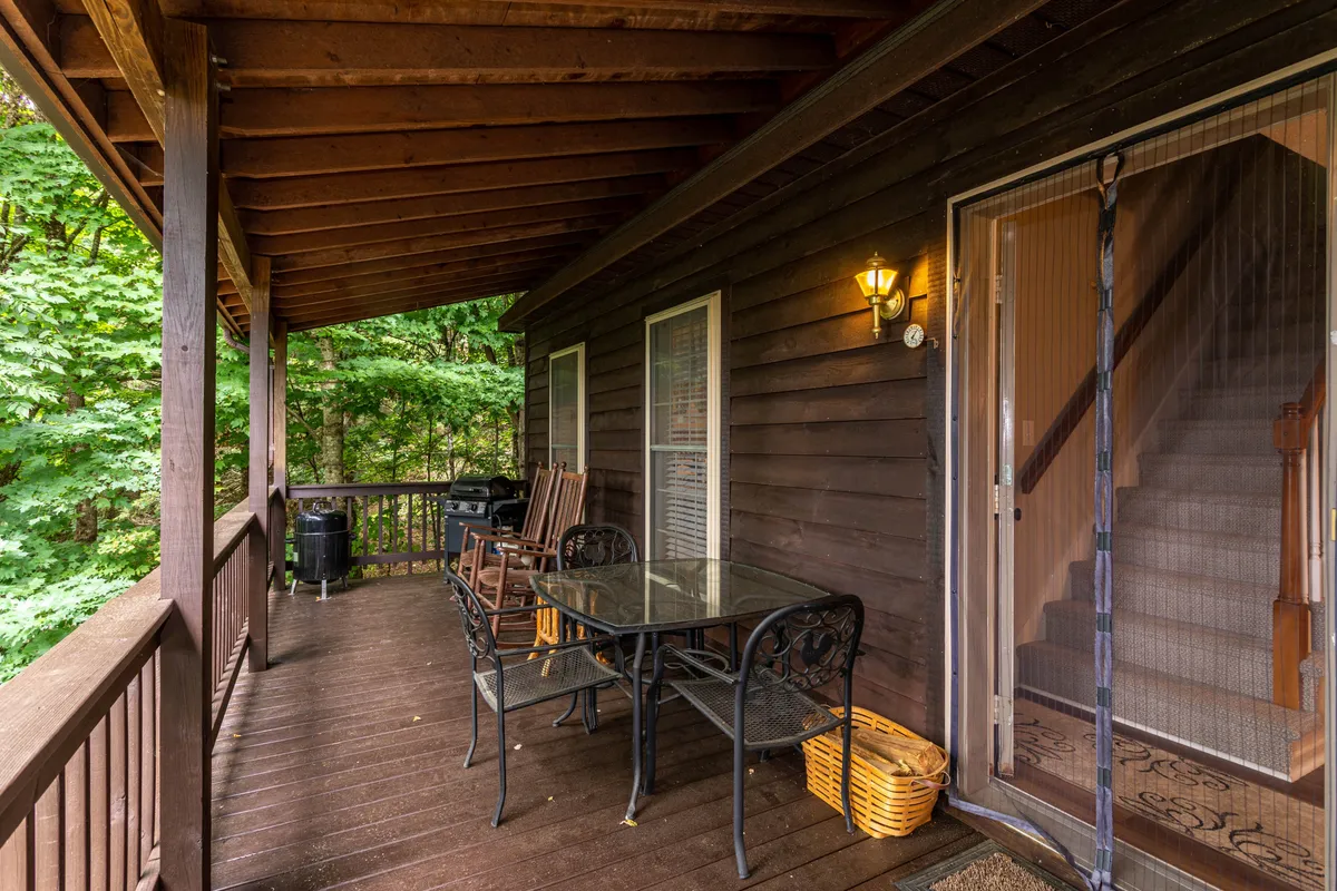 A welcoming deck scene with a glass table for outdoor dining, wooden rocking chairs for lounging, and lush greenery all around. The staircase and seating arrangement suggest this is a sociable space, perfect for sharing meals or relaxing after a day exploring the mountains.
