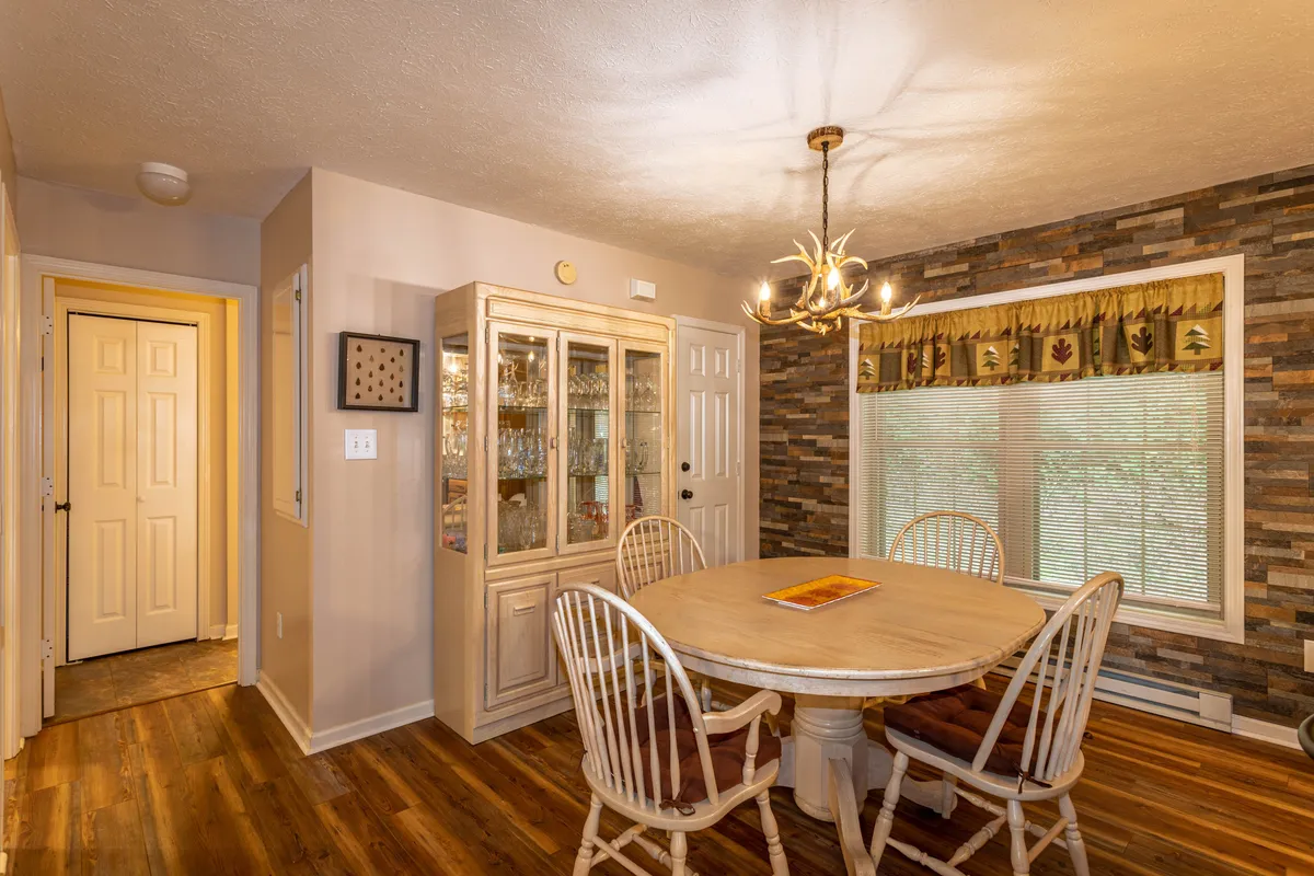 A warm and welcoming dining area featuring a wood table, glass-fronted cabinet, elegant chandelier, and a stone accent wall. The window with blinds lets in natural light, suggesting a connection to the outdoors and communal gathering after a day of adventure.