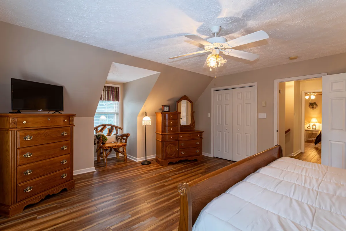 A bright and inviting bedroom with a wooden dresser, bed with crisp white bedding, TV, lamps, and a window offering natural light. The space feels both warm and modern, catering to relaxation and entertainment.