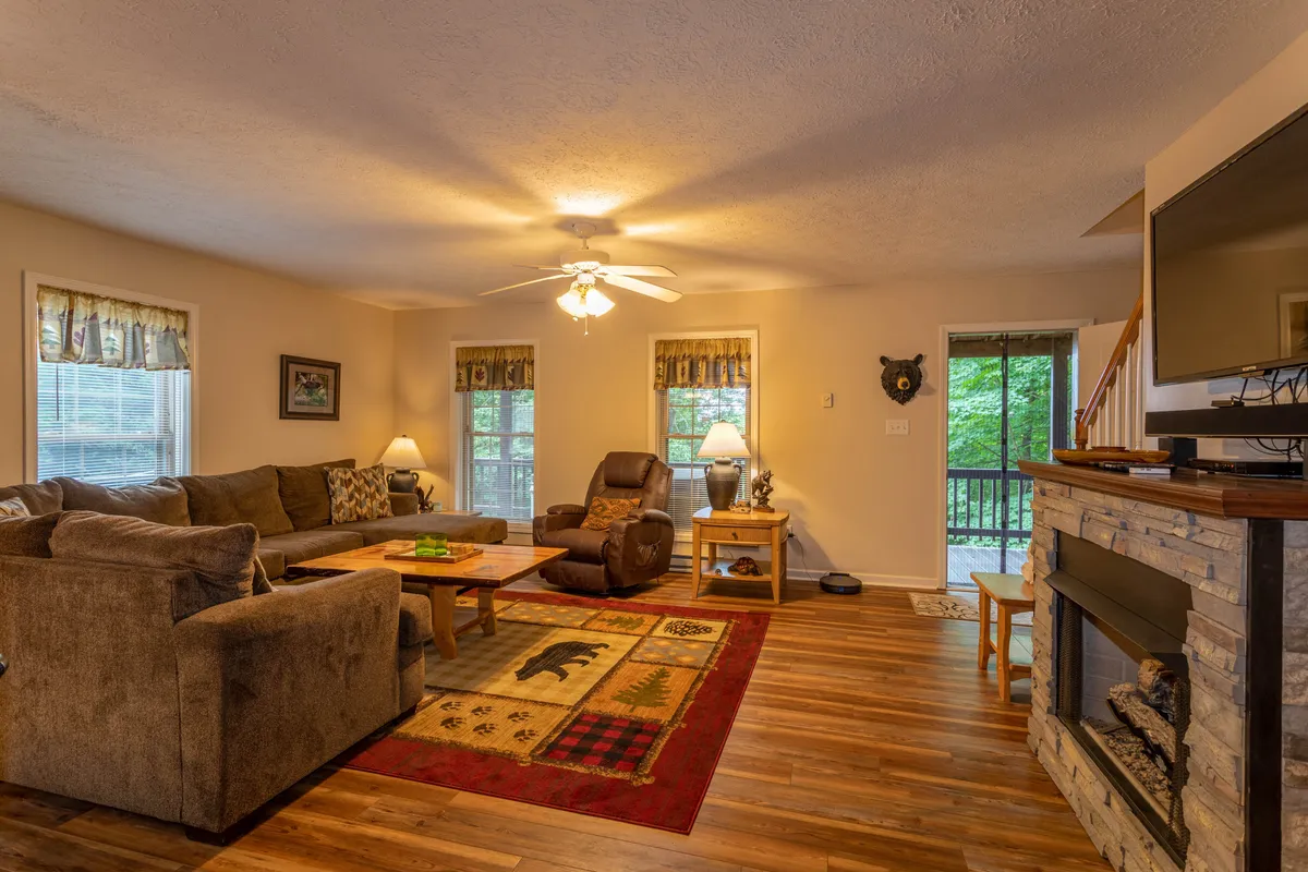 This image features a cozy living area with a plush sofa, wooden coffee table, a prominent stone fireplace, and large windows letting in natural light. The setting is perfect for relaxing or gathering with family and friends, with a warm and comfortable atmosphere that conveys the essence of mountain cabin living.