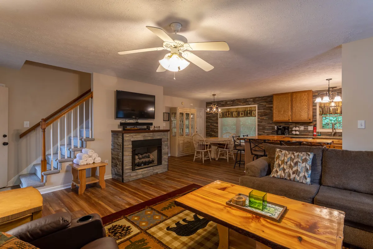 A warm, inviting living room with a large fireplace at its center, a wooden coffee table, and a view toward the dining area. The cozy space is perfect for family gatherings with a soft, welcoming ambiance, ideal for movie nights or relaxing after a day of exploration.