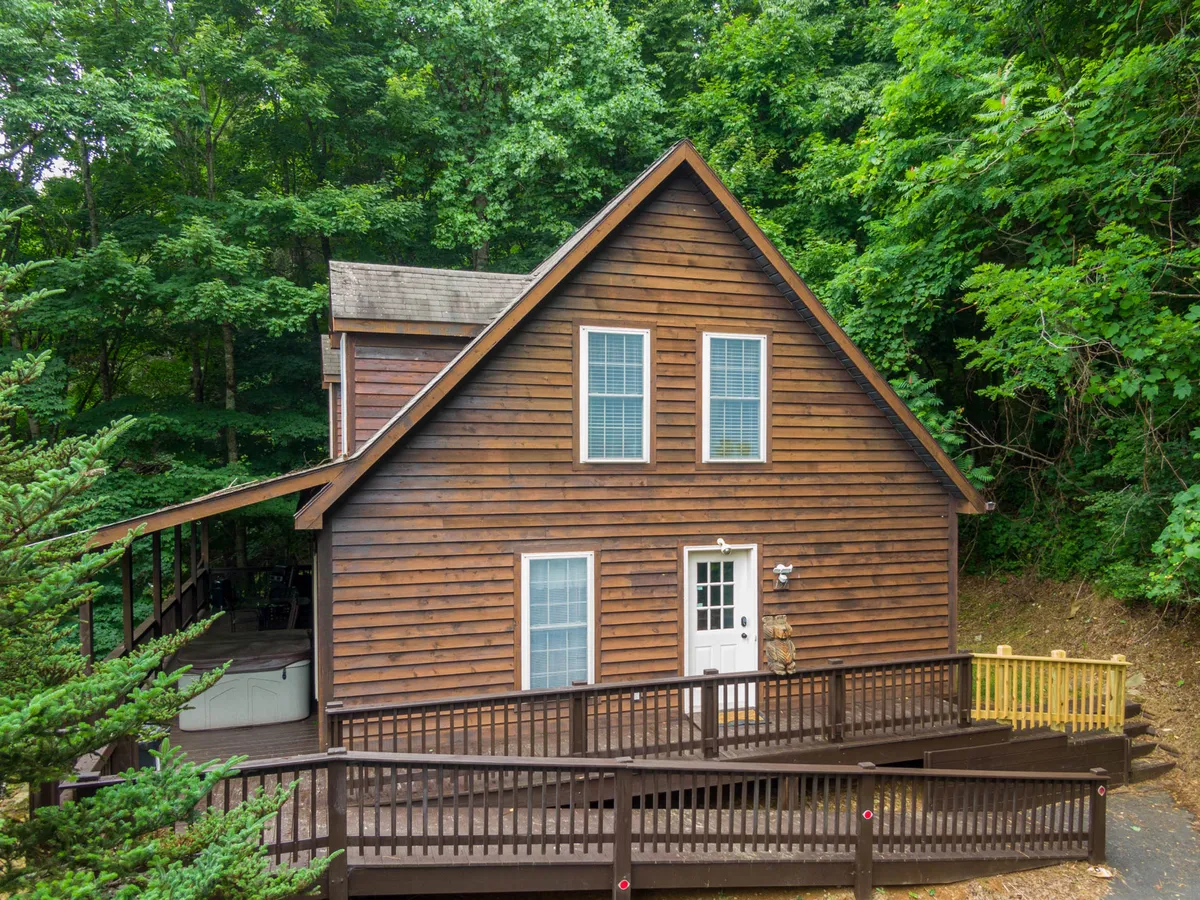 An exterior view of the cabin shows the wooden façade, inviting deck, and lush greenery surrounding the entryway. The deck offers space for outdoor gatherings, and the step-free front door suggests easy, accessible entry for all guests.
