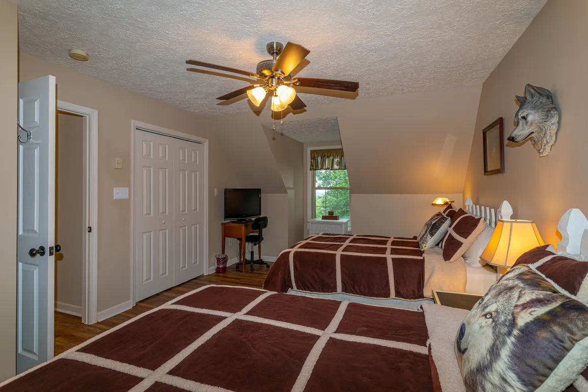 A bright bedroom with two beds adorned in brown plaid bedding, rustic wolf decor, a ceiling fan overhead, and a large window offering a view of surrounding greenery. The window visually extends the space toward the outdoors, evoking morning routines with pets before heading out to explore.