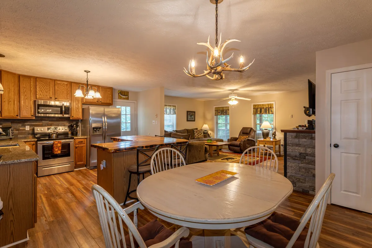 A spacious, open-plan dining and kitchen area that features a large table, kitchen island, and a comfortable sofa area nearby. The room is well-lit with a chandelier and is designed for shared meals, celebrations, and relaxed social time.